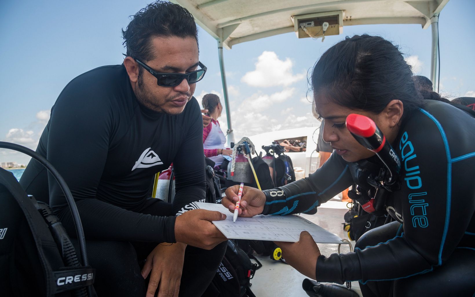 Two divers sit on a boat and work together over a paper on a clipboard.