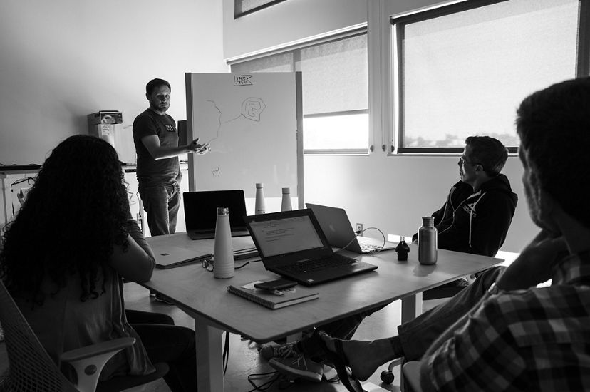A man stand next to a whiteboard, gesturing toward the simple drawing on it, while others look on