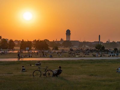 An old airport has been turned into green public space, with various people sitting on the grass, reading, and riding bikes.