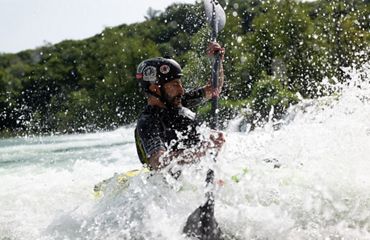 A kayaker wearing a helmet navigates through splashes and whitewater rapids on a sunny day.