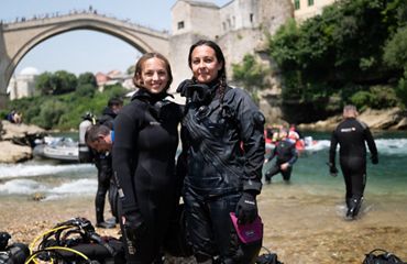 Two divers in wet suits smile and pose on a river bank with an old arched stone bridge in the background.