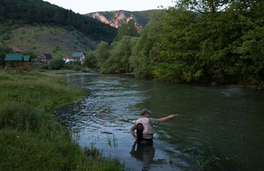A fisherman stands in a slow bend of a river and casts a rod around dusk with farms in the background.