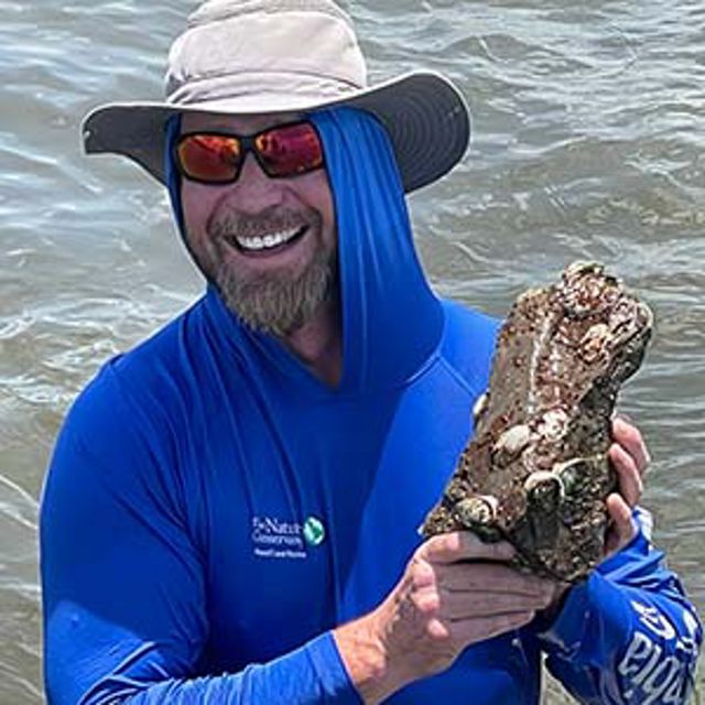 A man in waist-deep water holds up oysters on a lime rock while smiling.