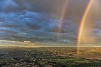 a wide view of rolling hills of developed land against a cloudy sky with two rainbows illuminated to the right of the shot.