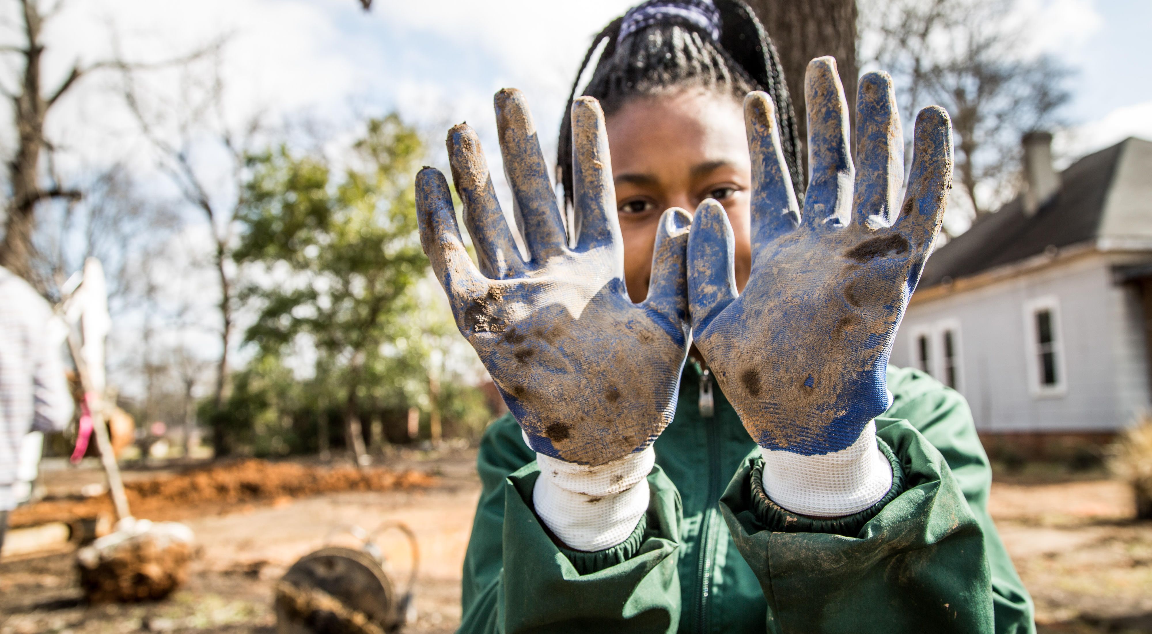 A student from historic Woodlawn High School in Birmingham, Alabama, shows off her dirty gloves from working on a "conservation lab." A "conservation lab" is vacant land converted into productive land for pollinators, birds, biodiversity, stormwater treatment demo areas, and generational care for the environment. These labs are low maintenance and a low-impact design.
