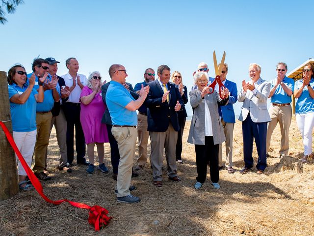 Several adults stand and applaud as Alabama Governor Kay Ivey holds up a giant pair of scissors after ceremoniously cutting a red ribbon to celebrate the completion of the Lightning Point restoration.