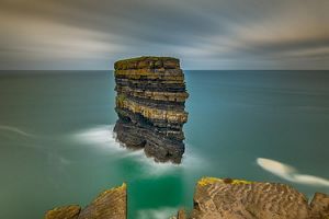 View from a high vantage point looking out at the green sea and a large rock formation composed of layers and layers of stacked rock.