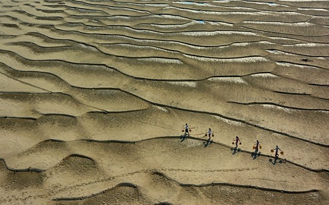 Aerial view of five people carrying water on their shoulders across a desert landscape.