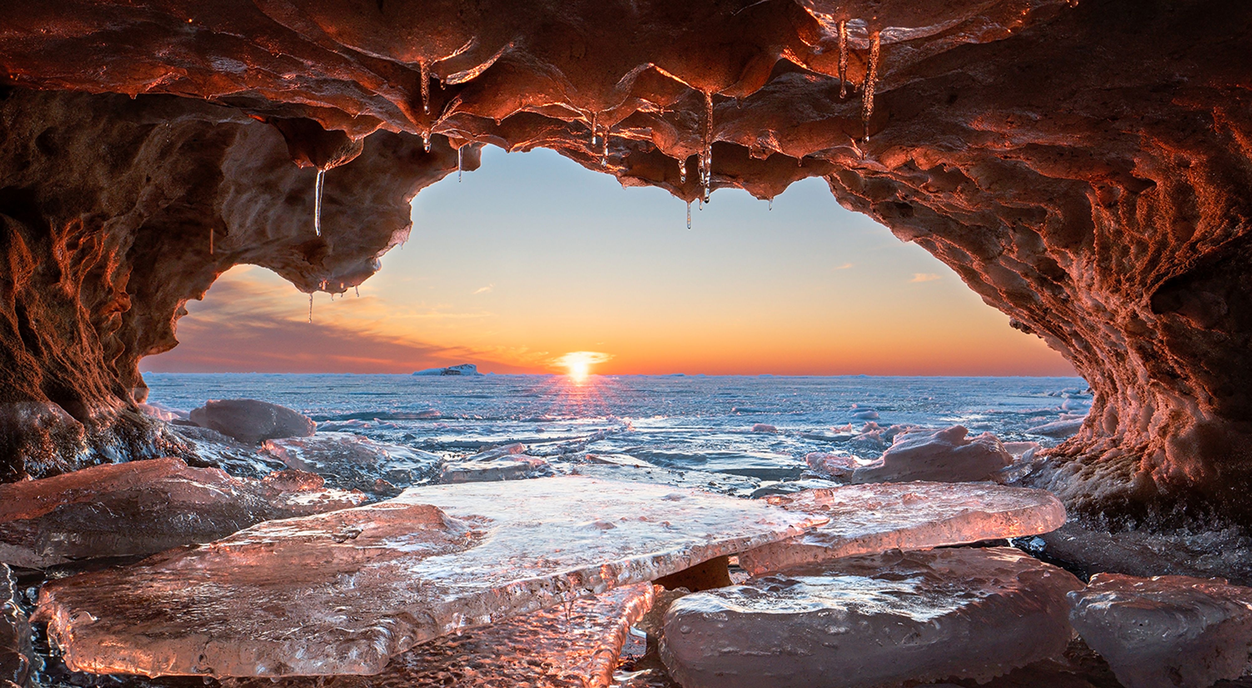 A small inlet within an ice shelf along the shoreline of Lake Michigan. Ice covers the ground as water rushes in. The sun sets on the horizon.