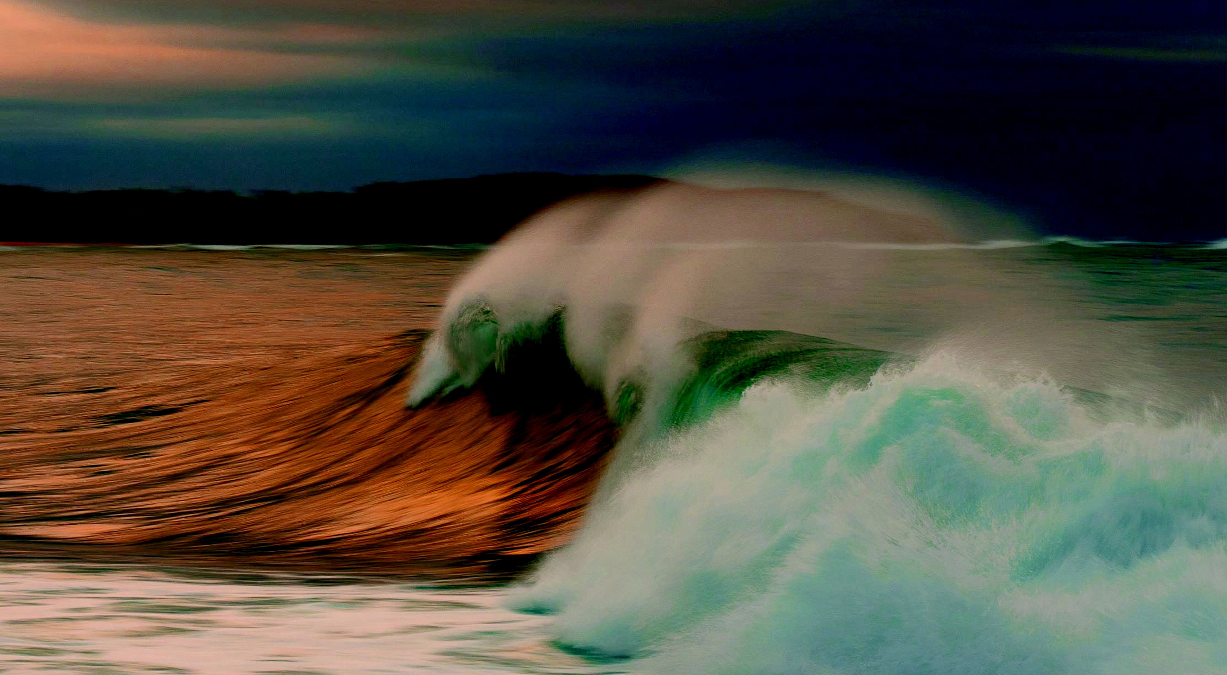 Photo of a wave crashing ashore under a dark sky.