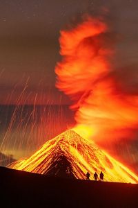  Nighttime view of a volcano erupting, with lava spewing up and flowing down the sides of the volcano and three silhouetted figures standing watching the eruption.