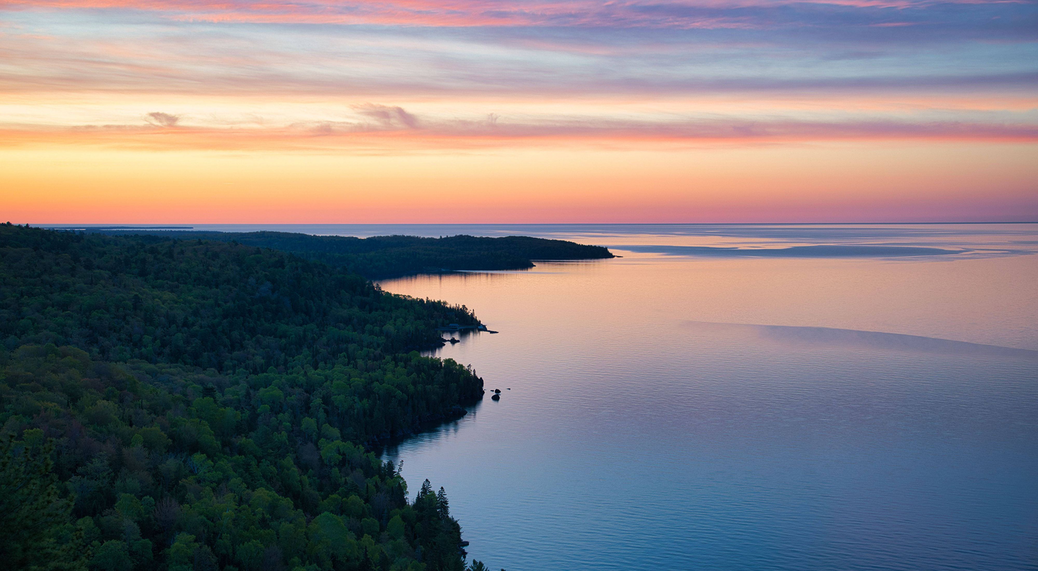 Colorful sunrise over Copper Harbor's peninsula in Michigan. The sky is filled with pastel pinks, oranges, and blues. The colors reflect in the water. The forest is thick with lush, green trees. 