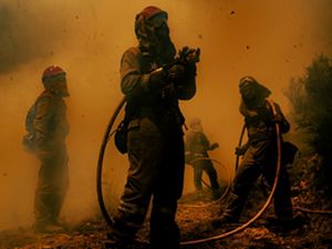 Four people in fire gear hold a water hose and stand at the edge of a forest. The air is orange and thick with smoke. Bits of ash and debris float in the air around them.