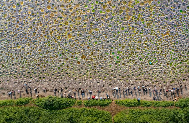 A group of people stand at the edge of a large, open sandy area. Large holes dot the surface, filled with green and orange water.