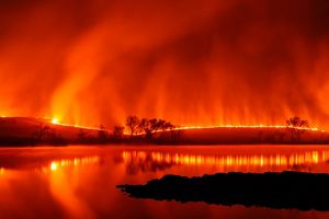 A glowing line of fire edges the horizon. Plumes of smoke rise in the orange tinged sky. The fire line is blurred and reflected in the surface of a lake.