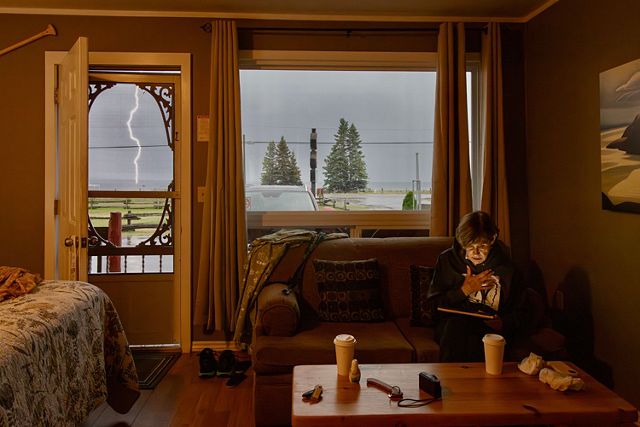 A woman sits on a sofa, her face softly illuminated by the glow of her tablet screen. A thick bolt of lightening is visible through the open door, splitting the gray sky.