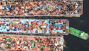 People and blankets create a brightly colored patchwork on the roof decks of three long boats. A smaller green decked boat floats at an angle behind the larger vessel in the center.
