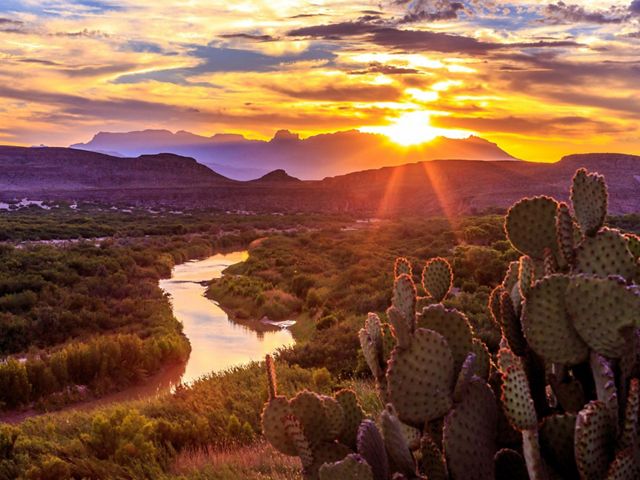 The sun sets over a winding river against a backdrop of mountains and cactuses.