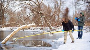 Brittney Rogers and Jacob Wojcik standing on a snowbank on the right side of the image. Brittney is holding a yellow pole with a 1-liter bottle attached to its end, dipping into the Salmon River.