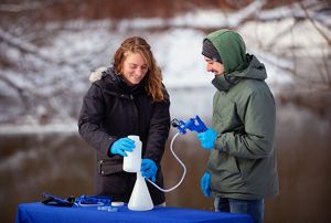Brittney Rogers and Jacob Wojcik operate different portions of testing equipment to examine whether eDNA is present in water samples. Both are wearing blue surgical gloves while doing so.