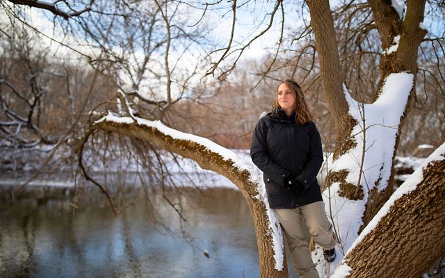 Brittney Rogers wears a black down coat and tan snow pants, leaning against a tree in the right hand side of the frame. She is looking off to the distance to the left.