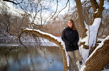 Brittney Rogers wears a black down coat and tan snow pants, leaning against a tree in the right hand side of the frame. She is looking off to the distance to the left.