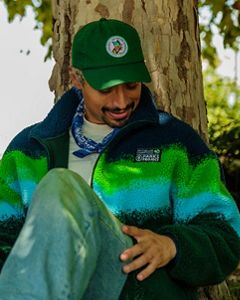 A man sits against the trunk of a birch tree looking at the Parks Project x TNC chest patch on his fleece jacket. His has short bleached hair and wears a green baseball style hat with the Parks Project x TNC 75th anniversary collab patch.