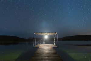 One person sits on a dock with an overhang while another person stands gazing up at brilliant stars shining against a dark sky.