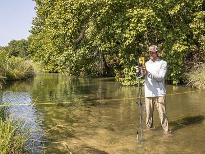 A man in a hat stands in a clear river lined with trees holding a tool to measure flow speed.