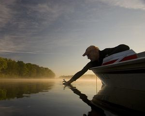 A man reaches out of a small boat and dips a glass bottle into the water.