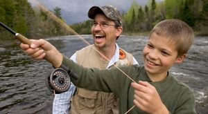 An adult and a child smile as they fly-fish on a wide river.