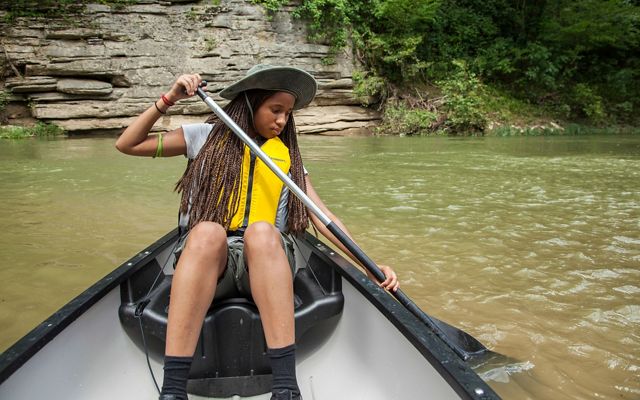 A TNC high school intern paddles a canoe on the green river with a rocky outcrop on the riverbank behind her.