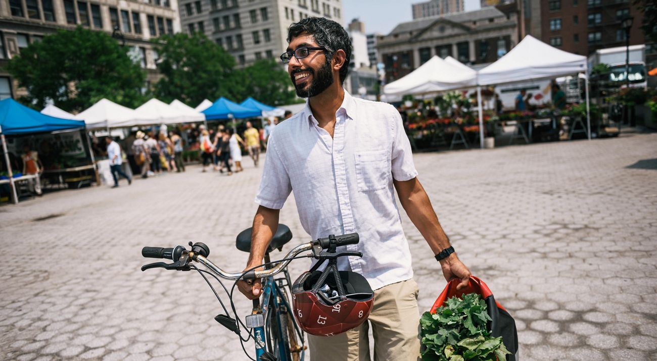 A smiling man walk a bike through an open plaza ringed by vendor tents. He is carrying a small tote filled with leafy greens.