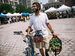 A man walks next to his bicycle in an urban farmers market; he carries a bag with leafy produce in it.