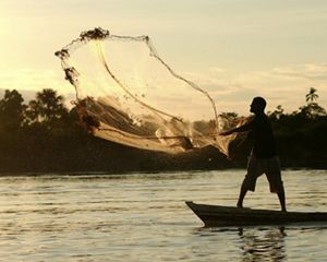 Silhueta de uma pessoa em pé em um barco lançando uma rede de pesca, iluminada por trás pelo sol poente.