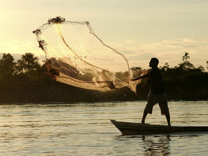 Pescador sobre canoa lanza una red de pesca en un rio.