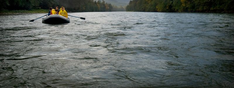 A group of people row a rubber raft along a wide river.