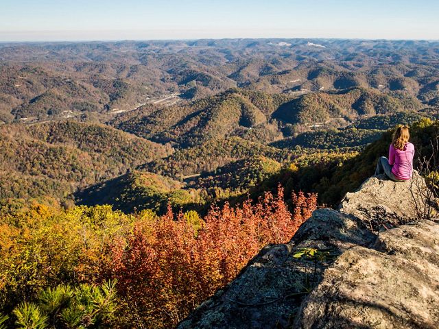 A hiker sits on a rock outcropping and looks out over rolling hills.