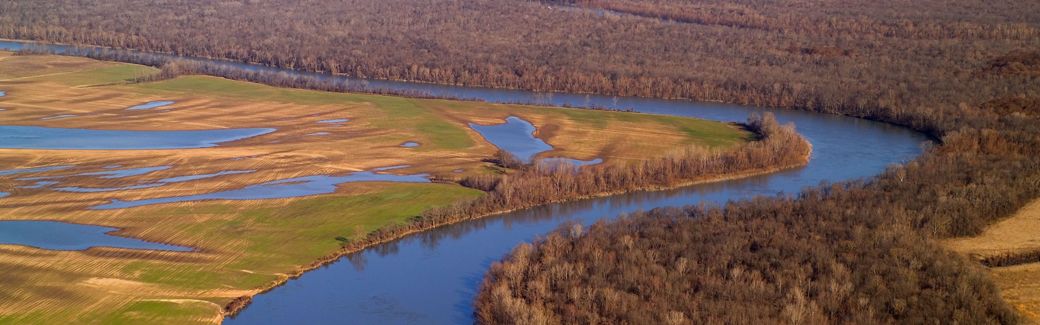 A bend in a river with fields on the left shore and forests on the right.