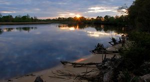 The sun sets over a placid body of water as a person stands on the water's banks and takes a photo.