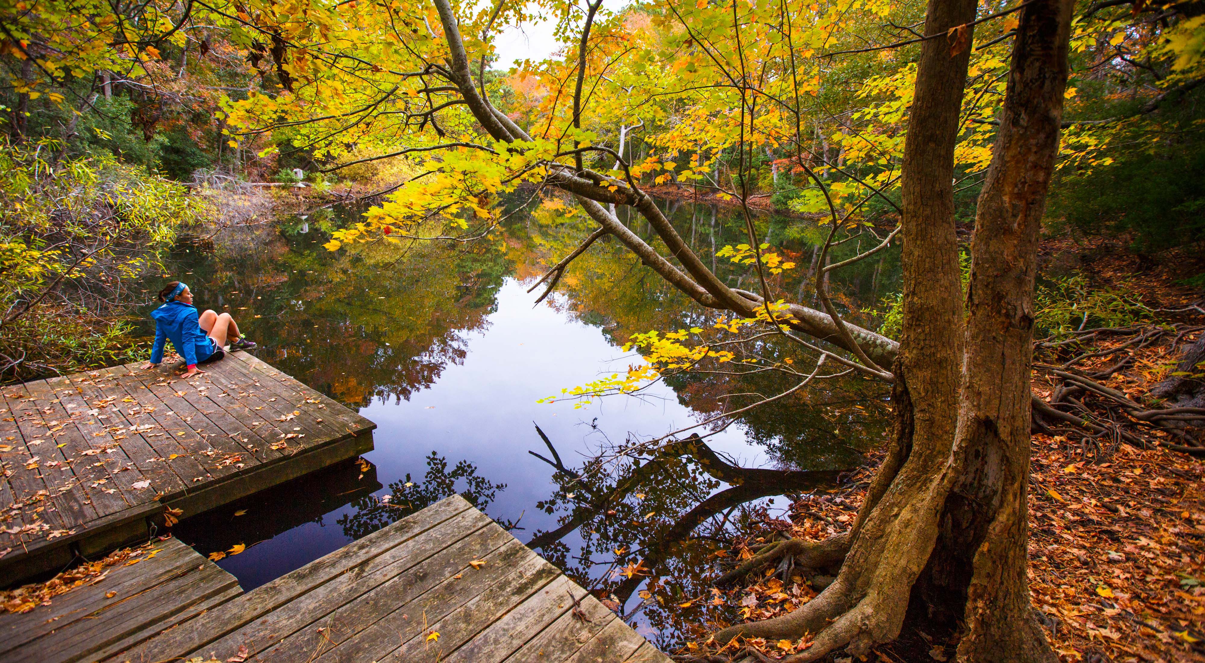 A young woman sits on a pier overlooking a pond, surrounded by trees with fall foliage.