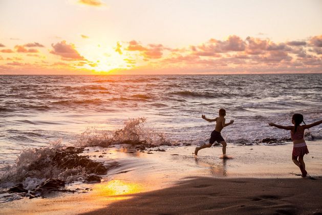 Two children play on the shoreline as a small waves crashes during sunset. 