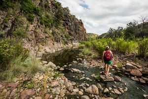 A woman with a backpack walks on stones in a river surrounded by mountains.