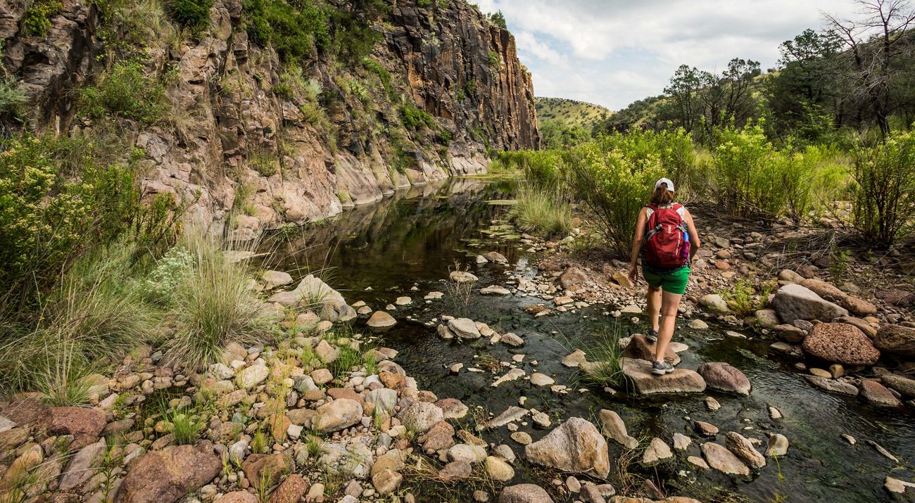 A woman with a backpack walks on stones in a river surrounded by mountains.