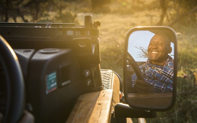 A smiling man's face is reflected in the side mirror of a Jeep being driven through the African bush.