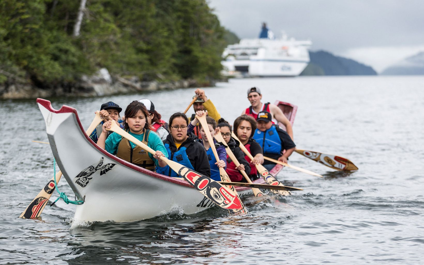 Several people paddle a traditional canoe on open water, with a cruise ship in the distance.