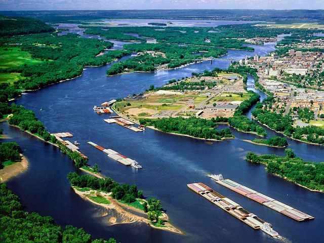 Vista aérea de barcaças no rio Mississippi e suas curvas contornando áreas povoadas e florestas.