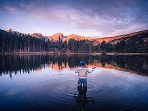 A man fishing in a lake surrounded by forest with mountains in the distance.