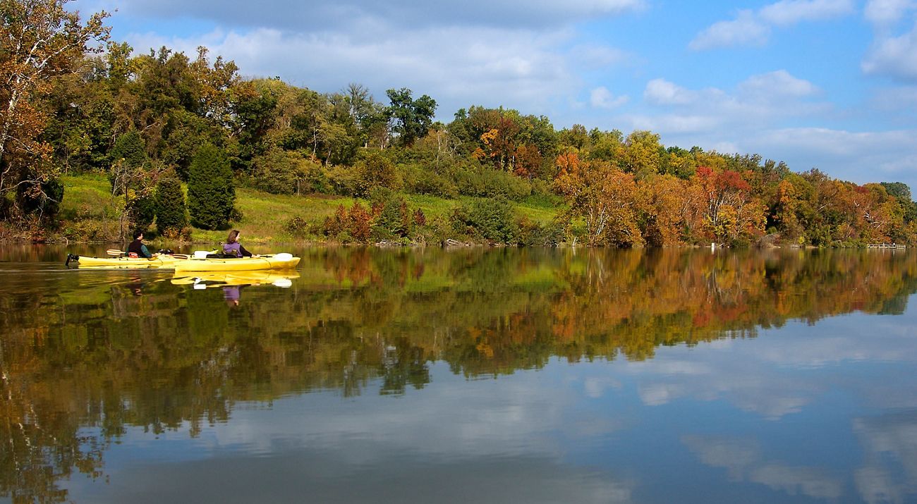 Two people in yellow kayaks float together down Nanjemoy Creek. The smooth, flat water reflects the autumn colors of the trees that line the creek bank.