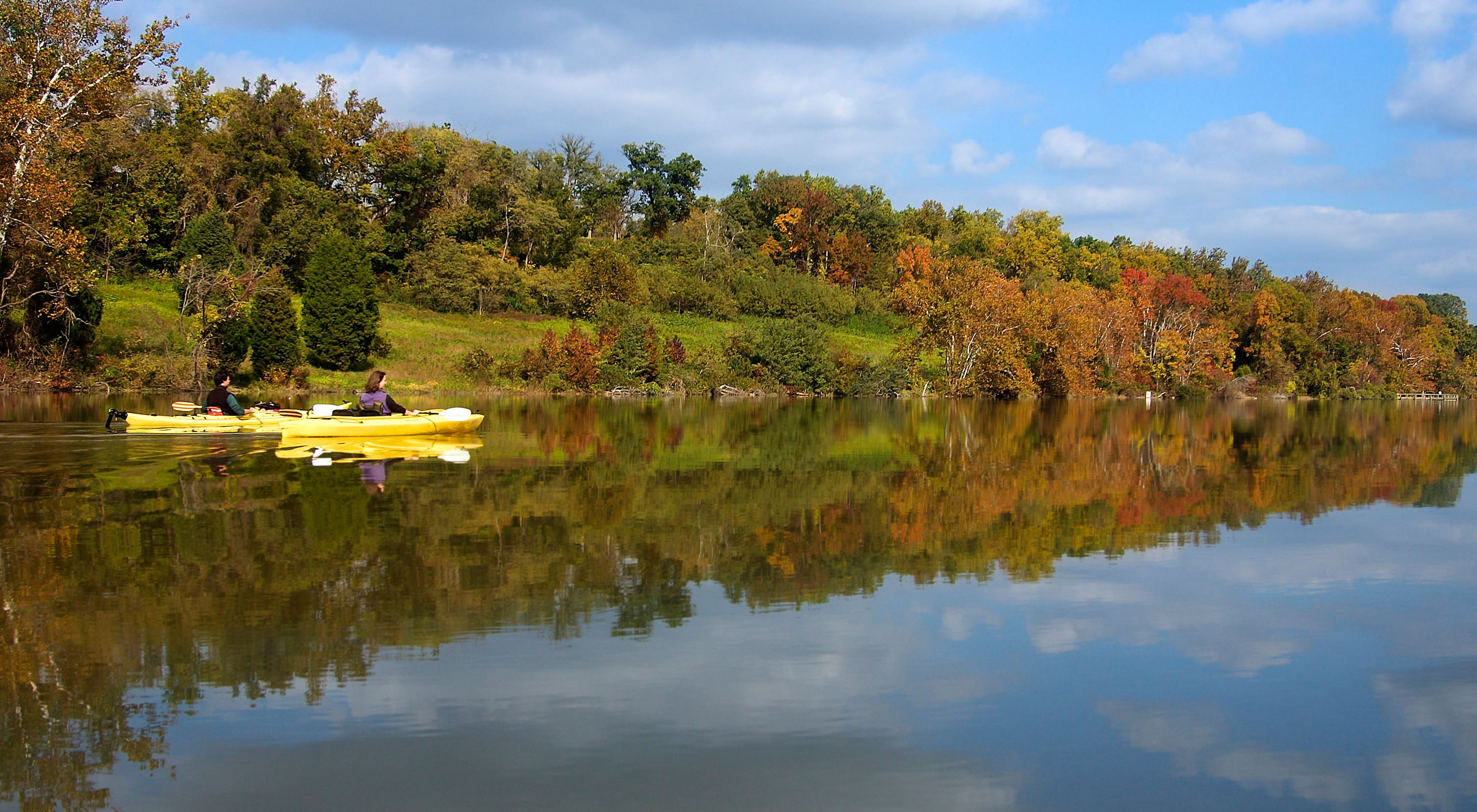 Two people in yellow kayaks float together down Nanjemoy Creek. The smooth, flat water reflects the autumn colors of the trees that line the creek bank.